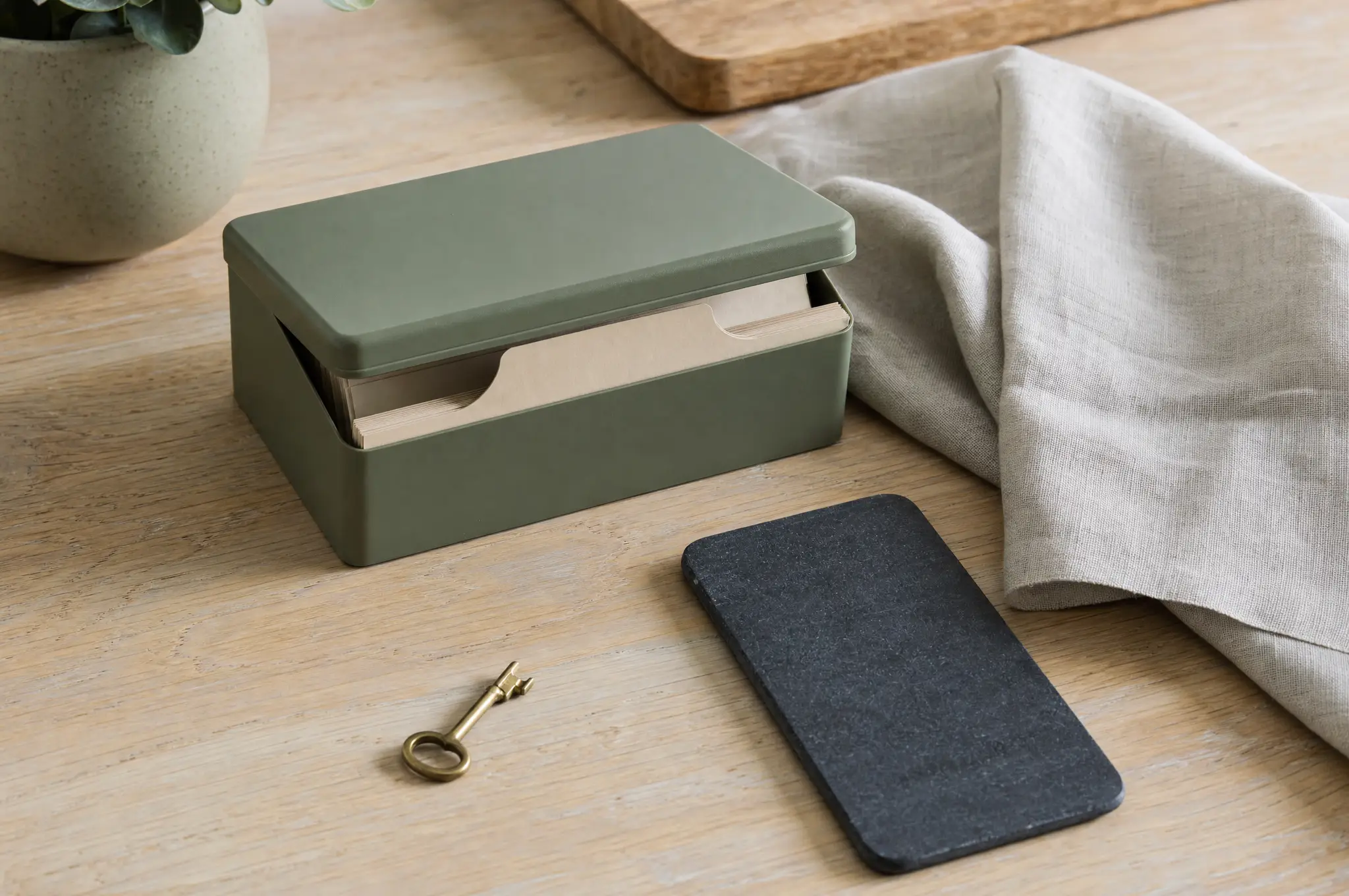 A closed personal recipe box with a source card, small key, and phone-shaped slate on a quiet kitchen table.